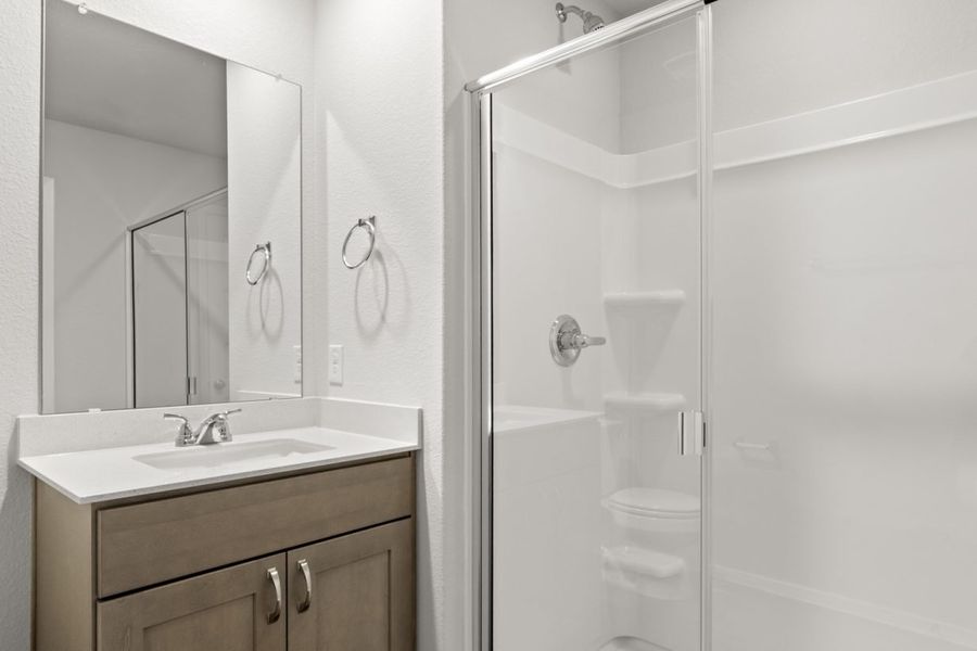 Image of a primary bathroom with a white vinyl walk-in shower, brown cabinets, and a white vanity with a mirror Image of a primary bathroom with a white vinyl walk-in shower, brown cabinets, and a white vanity with a mirror