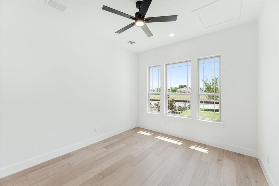 Empty bedroom with light wood floors, black ceiling fan, and three windows with backyard view