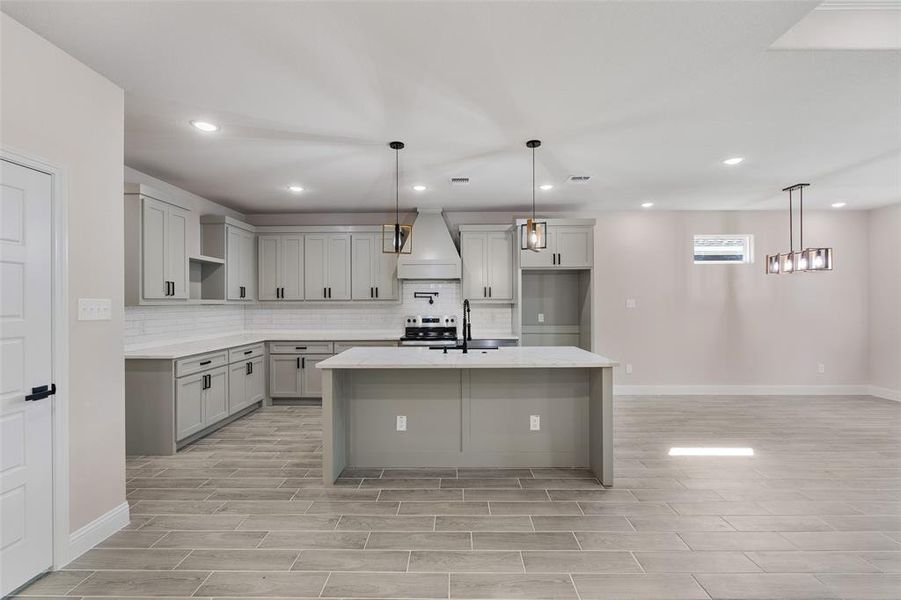 Kitchen featuring stainless steel range oven, custom range hood, an island with sink, decorative light fixtures, and tasteful backsplash