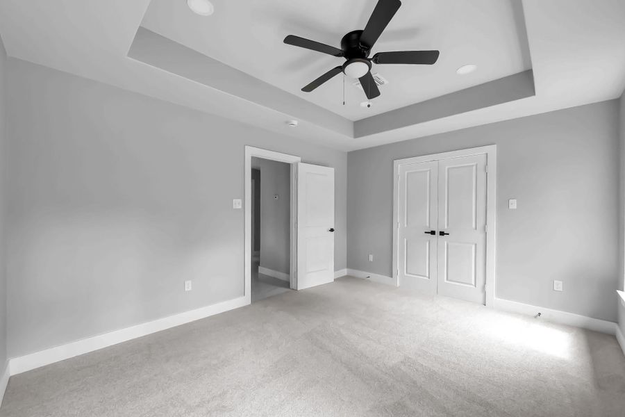 This spacious bedroom features neutral gray walls, plush carpet, and a ceiling fan with a tray ceiling design. It includes a double-door closet and receives ample natural light from a large window, creating a bright and airy atmosphere. This spacious bedroom features neutral gray walls, plush carpet, and a ceiling fan with a tray ceiling design. It includes a double-door closet and receives ample natural light from a large window, creating a bright and airy atmosphere.