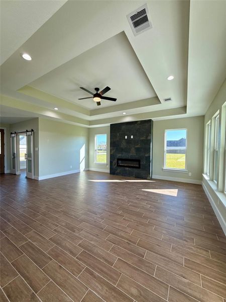 Spacious living room with wood-look tile flooring, modern recessed lighting, and a unique tray ceiling. Features a sleek fireplace, large windows for ample natural light, and a ceiling fan. Ideal for contemporary living. Spacious living room with wood-look tile flooring, modern recessed lighting, and a unique tray ceiling. Features a sleek fireplace, large windows for ample natural light, and a ceiling fan. Ideal for contemporary living.