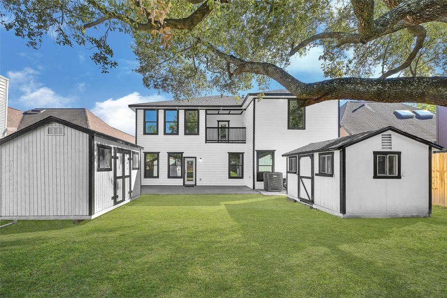 This photo showcases a modern two-story home with a spacious backyard, featuring two small outbuildings. The yard is shaded by a large tree, providing a serene outdoor space. The house has a clean, white exterior with large windows, allowing for ample natural light.