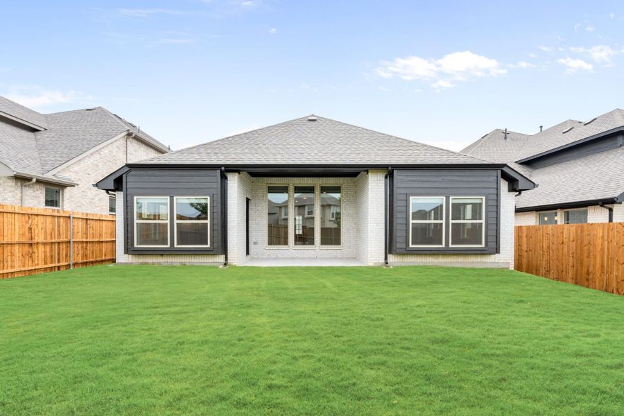 Rear exterior of single-story home with gray siding, brick accents, patio, and fenced backyard with green lawn Rear exterior of single-story home with gray siding, brick accents, patio, and fenced backyard with green lawn