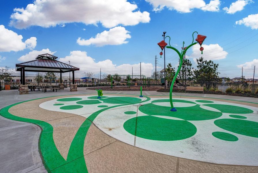This photo showcases a community park area featuring a splash pad with playful water features and a covered pavilion with picnic tables, ideal for family gatherings and outdoor activities. The setting is bright and open, offering a welcoming environment for residents.