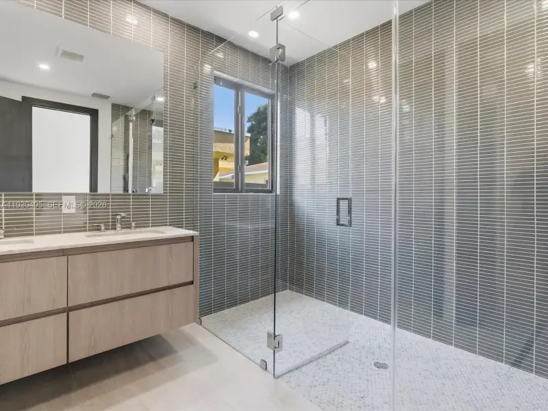 Floating vanity with light wood-finish cabinetry, quartz countertop, floor-to-ceiling vertically stacked tile, frameless glass shower enclosure, and circular mosaic floor tile Floating vanity with light wood-finish cabinetry, quartz countertop, floor-to-ceiling vertically stacked tile, frameless glass shower enclosure, and circular mosaic floor tile