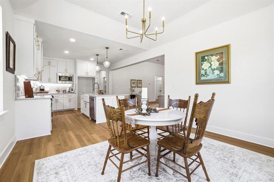Open-concept dining area featuring wood-finish flooring, a contemporary chandelier, and a sightline to the kitchen with white cabinetry, stainless steel appliances, and a central island with pendant lighting Open-concept dining area featuring wood-finish flooring, a contemporary chandelier, and a sightline to the kitchen with white cabinetry, stainless steel appliances, and a central island with pendant lighting