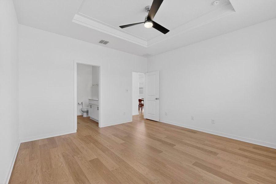 Unfurnished bedroom featuring a ceiling fan, ensuite bathroom, light wood-type flooring, and a tray ceiling