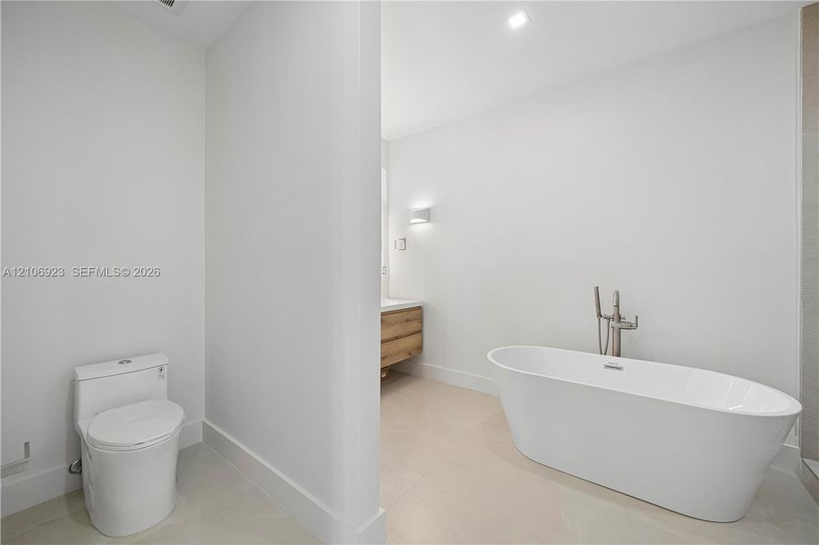 Freestanding white soaking tub with polished chrome floor-mounted faucet, wood-finish vanity with countertop basin, and low-profile elongated toilet Freestanding white soaking tub with polished chrome floor-mounted faucet, wood-finish vanity with countertop basin, and low-profile elongated toilet