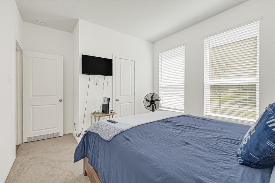 Bright and cozy bedroom featuring large windows with blinds, a comfortable bed with blue bedding, carpeted flooring, and a wall-mounted TV. Bright and cozy bedroom featuring large windows with blinds, a comfortable bed with blue bedding, carpeted flooring, and a wall-mounted TV.