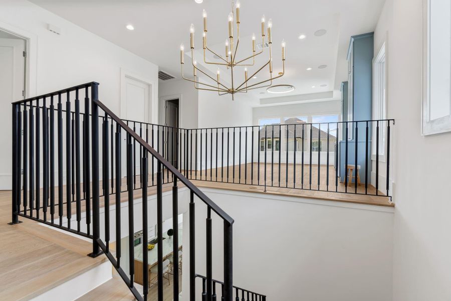The upstairs landing overlooks the main living area below, featuring modern iron railings and a statement chandelier that enhances the home’s open, airy feel.