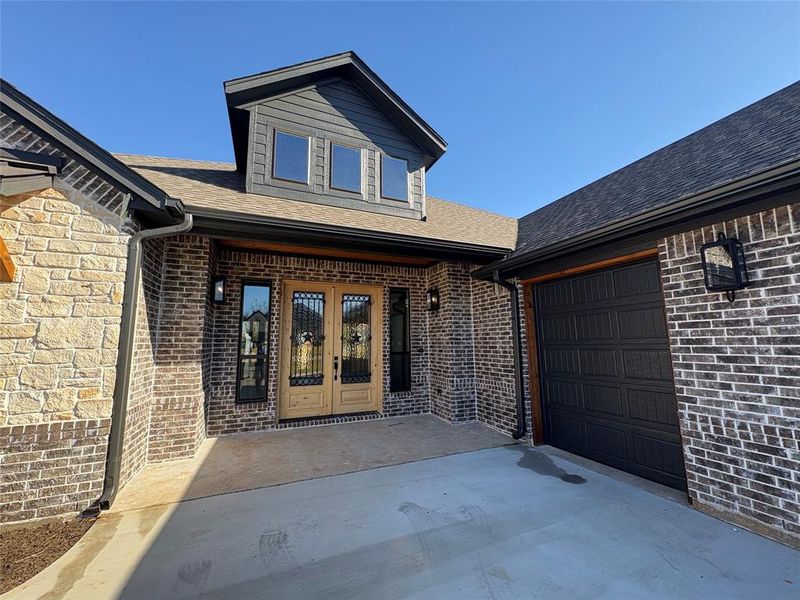 Doorway to property featuring a shingled roof, french doors, brick siding, a garage, and a patio area
