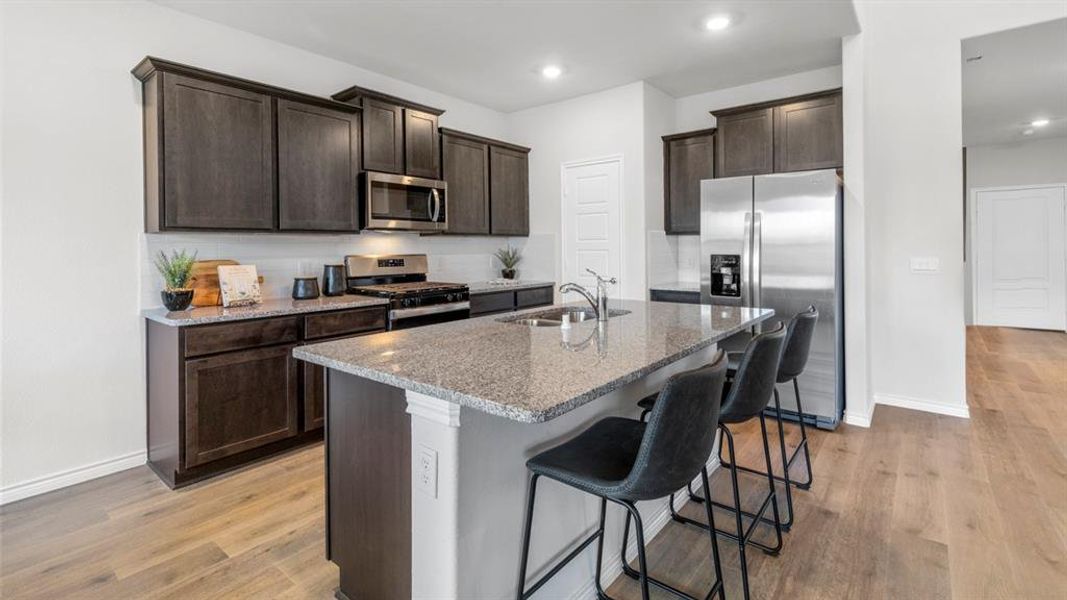 Kitchen featuring an island with seating, dark wood cabinetry, stainless steel appliances, speckled countertops, and wood-finish flooring Kitchen featuring an island with seating, dark wood cabinetry, stainless steel appliances, speckled countertops, and wood-finish flooring
