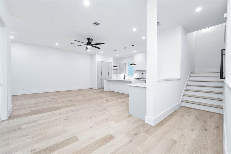 A modern and spacious open-concept living area with fresh white walls, light wood flooring, contemporary ceiling fans, and a sleek kitchen with an island. The space is well-lit with recessed lighting and features a staircase leading to another level.