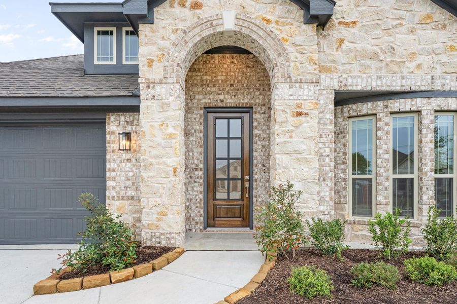 Stone and brick home exterior with arched entryway, wood front door with glass panels, and gray garage door Stone and brick home exterior with arched entryway, wood front door with glass panels, and gray garage door