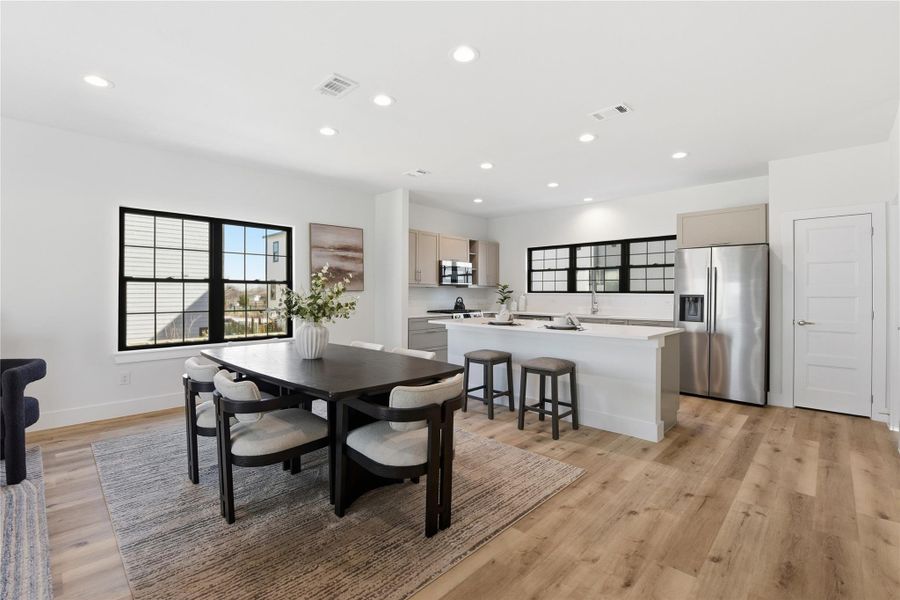 A modern open-concept kitchen and dining area with hardwood floors, white walls, recessed lighting, a spacious island, and large windows that fill the space with natural light.