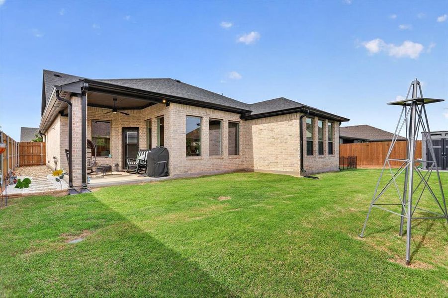 Expansive backyard featuring a covered patio with a ceiling fan, a brick exterior, multiple windows with dark framing, and a wooden privacy fence