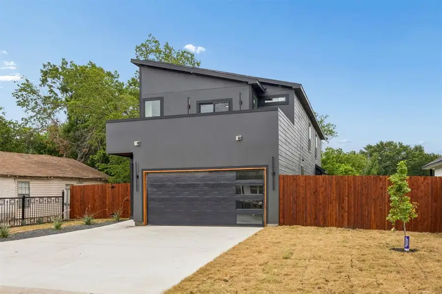 Modern architectural exterior featuring a dark gray facade, an attached two-car garage with frosted glass panels, a private wooden fence, a concrete driveway, and a sloped roofline Modern architectural exterior featuring a dark gray facade, an attached two-car garage with frosted glass panels, a private wooden fence, a concrete driveway, and a sloped roofline