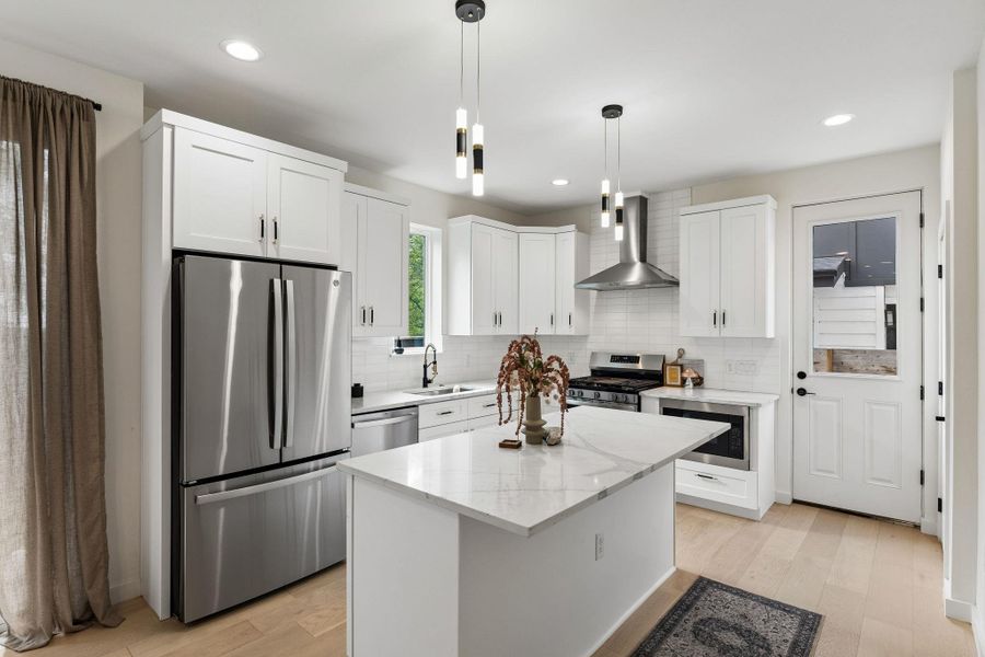 A full view of the kitchen showcasing the functional layout. The large center island with waterfall quartz countertops serves as a focal point, complemented by elegant pendant lighting. A French-door refrigerator and a glass-paneled back door add to the room’s bright and airy atmosphere.
