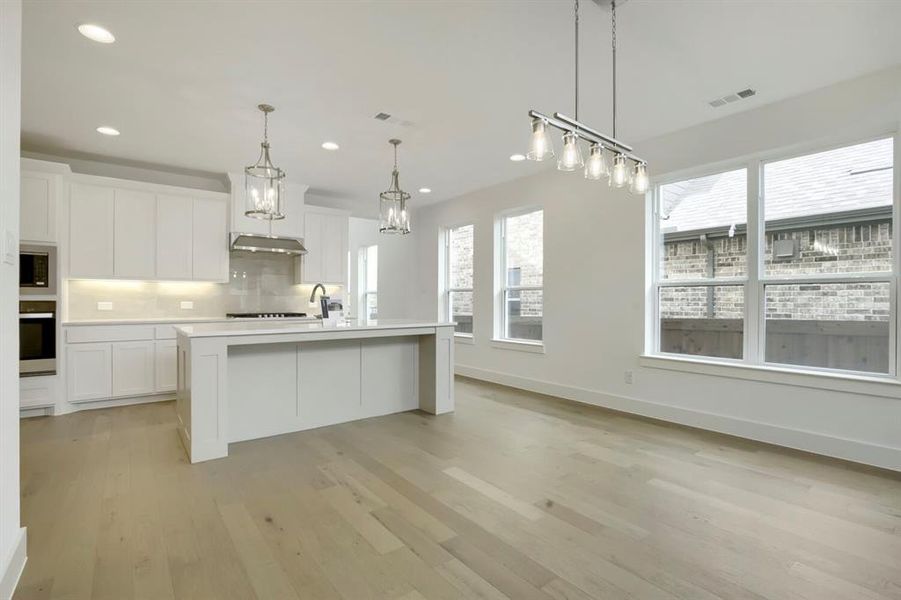 Open-concept kitchen and dining area featuring light wood-finish flooring, white cabinetry, a central island with countertop seating, and multiple windows providing ambient light