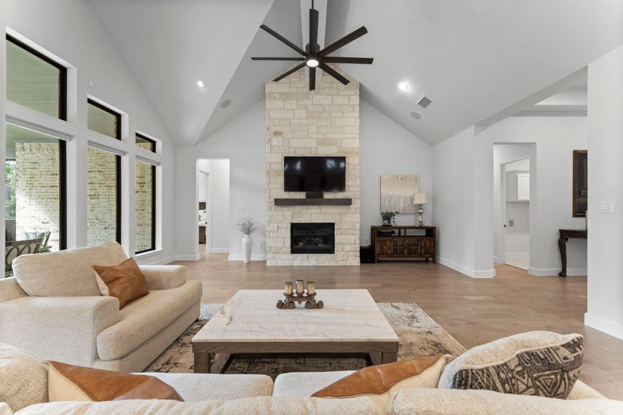 This image captures a spacious, open living room with a vaulted ceiling and light wood floors. The focal point is a floor-to-ceiling stone fireplace wooden mantel. Large windows and a glass door on the left provide a view of the covered patio. This image captures a spacious, open living room with a vaulted ceiling and light wood floors. The focal point is a floor-to-ceiling stone fireplace wooden mantel. Large windows and a glass door on the left provide a view of the covered patio.