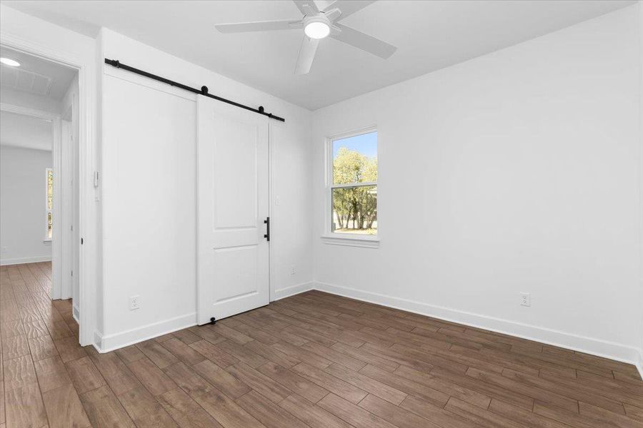 Unfurnished bedroom with a barn door, wood finished floors, baseboards, and multiple windows Unfurnished bedroom with a barn door, wood finished floors, baseboards, and multiple windows