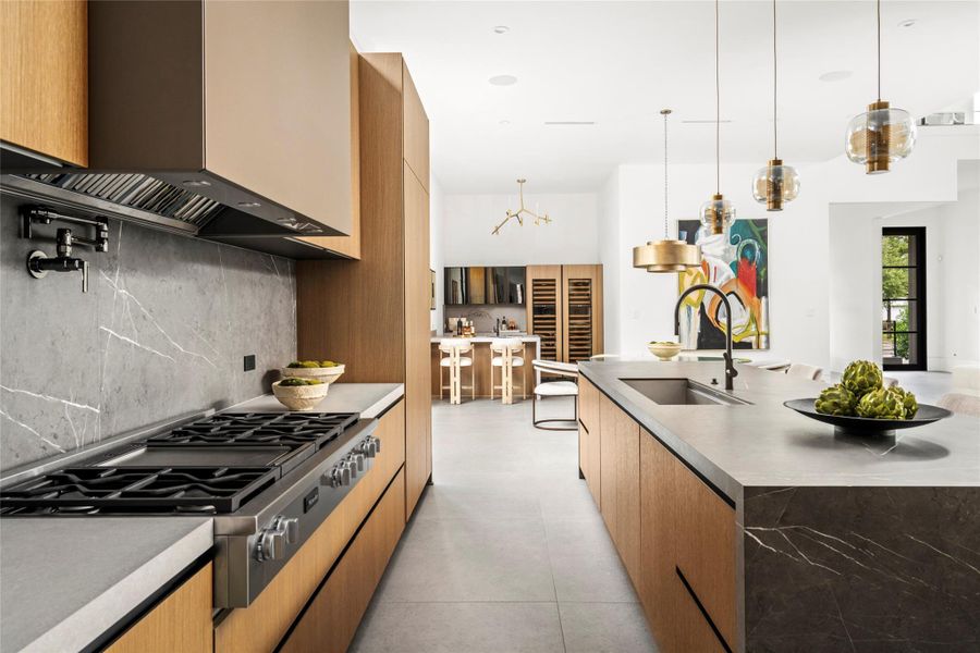 [Gathering Kitchen] Italian-engineered cabinetry with fully integrated appliance panels, concealed hardware, and continuous grain match across every run. Miele 48" gas range with griddle, pot filler, concealed vent hood, full-height stone slab backsplash, and waterfall island. Adjacent wet bar with climate-controlled wine storage.