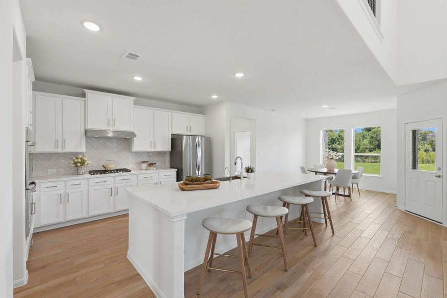 This photo showcases a modern, open-concept kitchen and dining area with white cabinetry, a large island with bar seating, stainless steel appliances, and wood-style flooring. Large windows provide abundant natural light and views of the greenery outside.