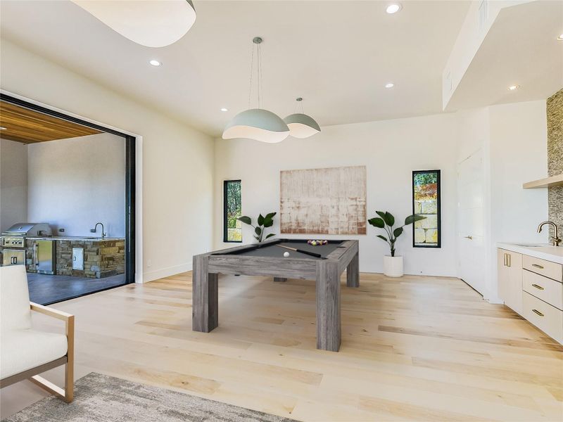 This versatile room features light-toned wood flooring, recessed lighting, and a wet bar with light cabinetry and a stone-tiled backsplash