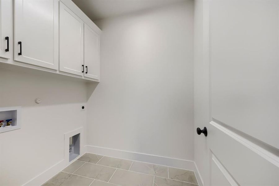 Utility room featuring upper cabinetry with black hardware, white walls, and light-toned rectangular floor tiles