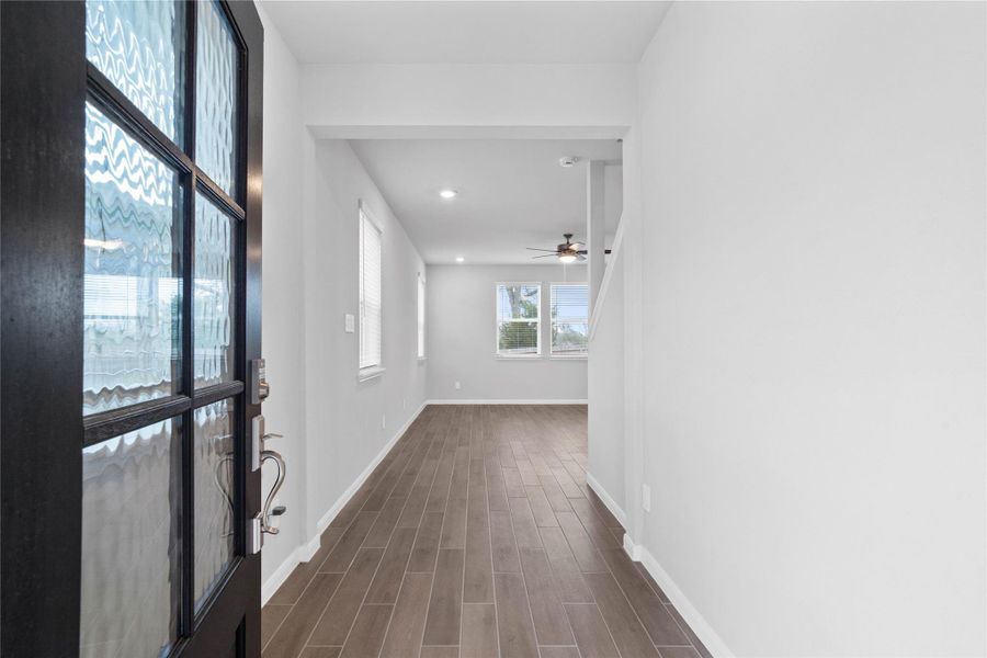 This entryway features a modern, open-concept design. The long, bright corridor utilizes a neutral color palette, recessed lighting, and wood-look tile flooring to lead the eye toward the home's main living area.