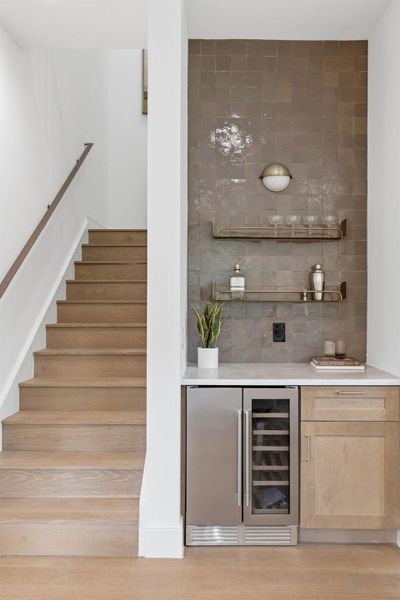 Elegant wet bar with a wine cooler and built-in shelving, complemented by rich hardwood flooring and a stylish staircase.