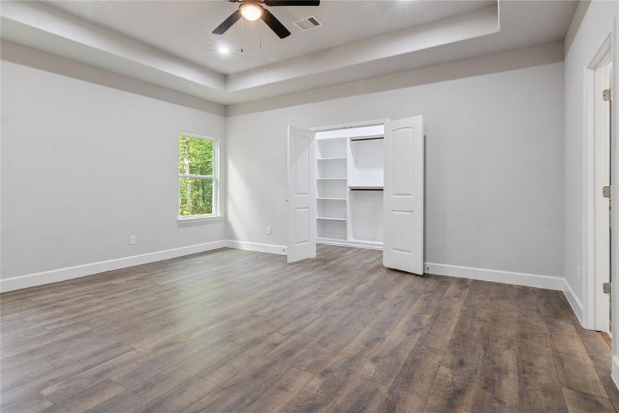 This spacious bedroom features beautiful floors, a tray ceiling with recessed lighting and a ceiling fan, a window offering natural light, and an open closet with ample storage.