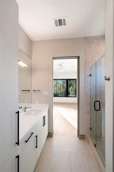 This photo showcases a modern bathroom featuring a sleek white vanity with black hardware, a large mirror, and a glass-enclosed shower. The room is bright, thanks to natural light from an adjacent room visible through the open door, offering views of greenery outside.