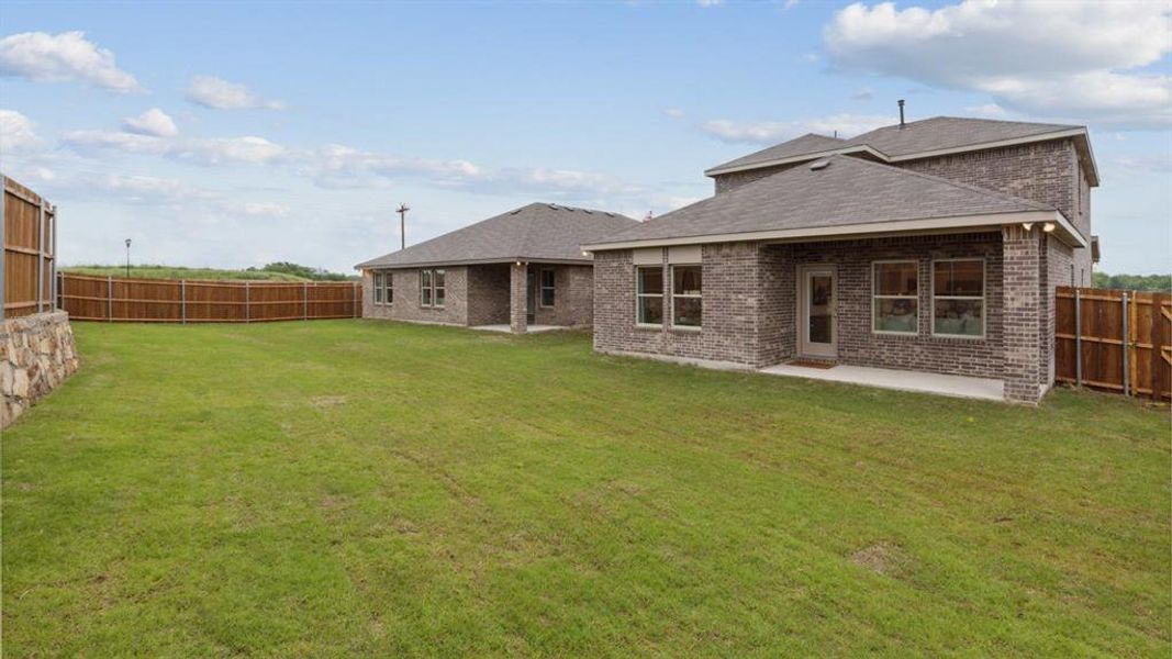 Expansive grassy backyard featuring a full privacy fence, stone retaining wall, and a covered concrete patio