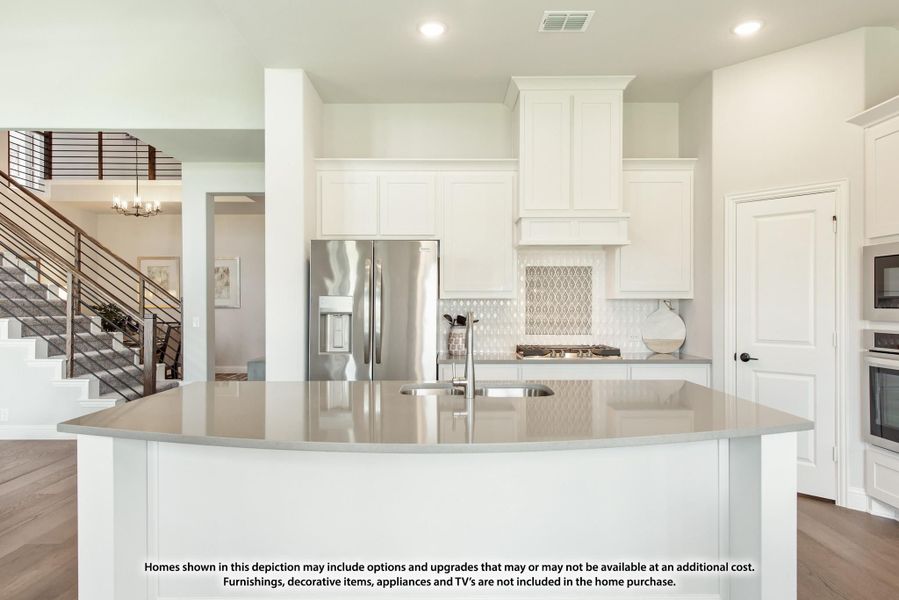 Kitchen with large curved island, white cabinets, stainless steel refrigerator, and patterned tile backsplash with staircase visible in background.