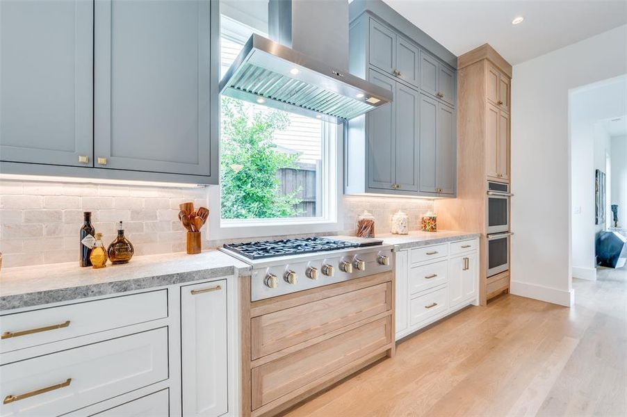 Kitchen with tasteful backsplash, wall chimney exhaust hood, light stone countertops, appliances with stainless steel finishes, and light hardwood / wood-style flooring