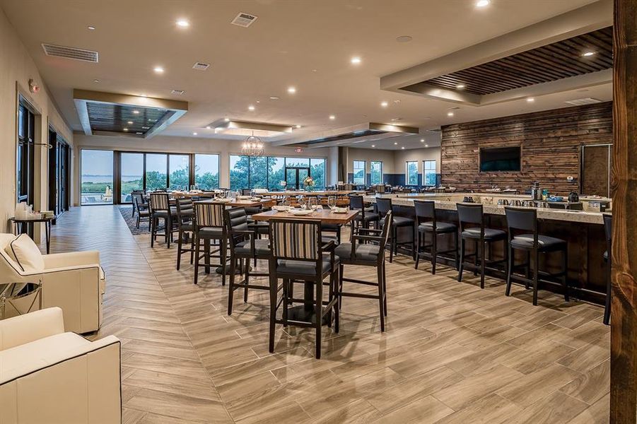 Spacious interior featuring a large bar with a stone countertop and wood-paneled accent wall, a dining area with multiple tables, wood-look plank flooring in a herringbone pattern, recessed lighting, and expansive glass doors opening to the exterior Spacious interior featuring a large bar with a stone countertop and wood-paneled accent wall, a dining area with multiple tables, wood-look plank flooring in a herringbone pattern, recessed lighting, and expansive glass doors opening to the exterior