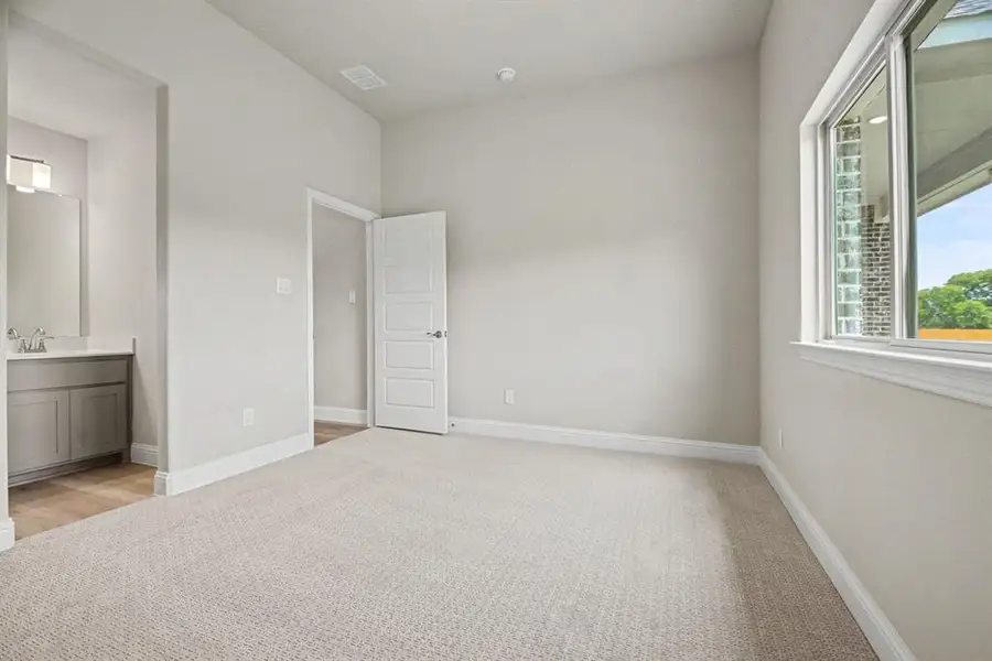 Neutral-toned interior featuring light gray walls, plush carpeting, a multi-pane window with white trim, a solid panel door, and a vanity area with a gray cabinet and rectangular mirror Neutral-toned interior featuring light gray walls, plush carpeting, a multi-pane window with white trim, a solid panel door, and a vanity area with a gray cabinet and rectangular mirror