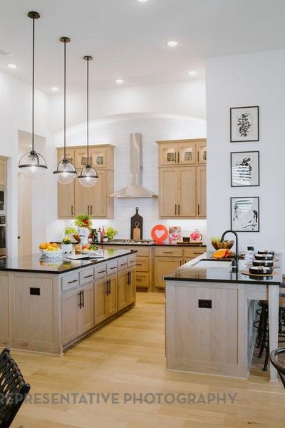 Kitchen with wall chimney range hood, dark countertops, light brown cabinetry, light wood-type flooring, and a center island Kitchen with wall chimney range hood, dark countertops, light brown cabinetry, light wood-type flooring, and a center island