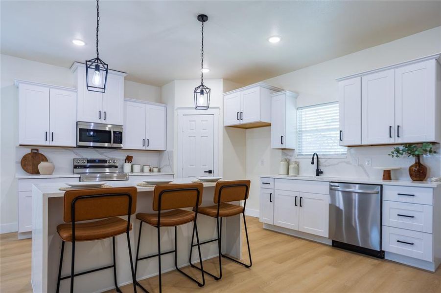 Kitchen with a breakfast bar area, appliances with stainless steel finishes, white cabinetry, an island with sink, and hanging light fixtures