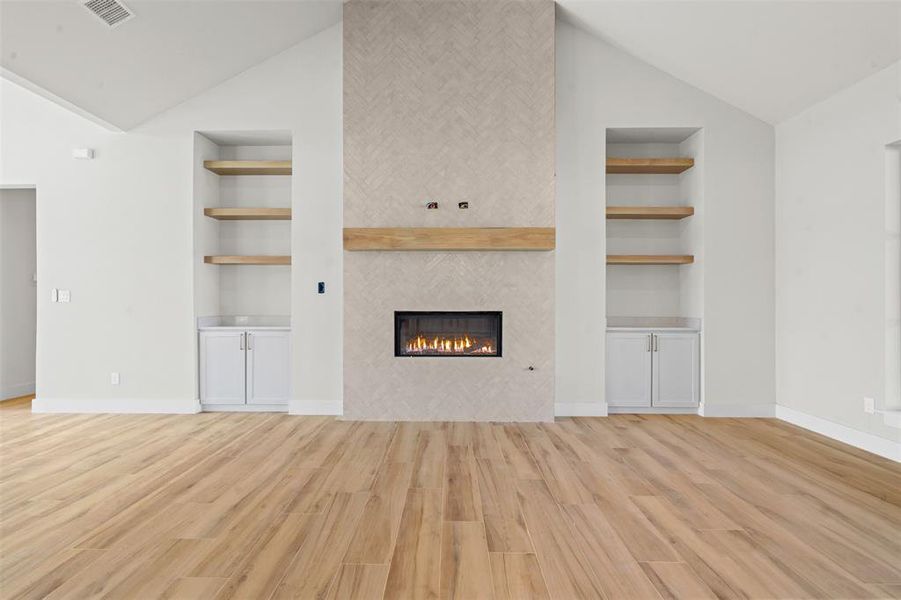 Unfurnished living room with built in shelves, lofted ceiling, light wood-style flooring, and a tiled fireplace Unfurnished living room with built in shelves, lofted ceiling, light wood-style flooring, and a tiled fireplace