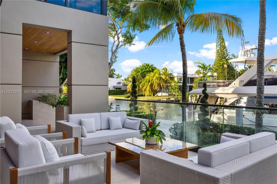 Ground-level waterfront terrace with glass railing, woven outdoor sectional and lounge seating, direct canal views, private dock with yacht, tropical palm landscaping, and wood-ceiling loggia beyond.