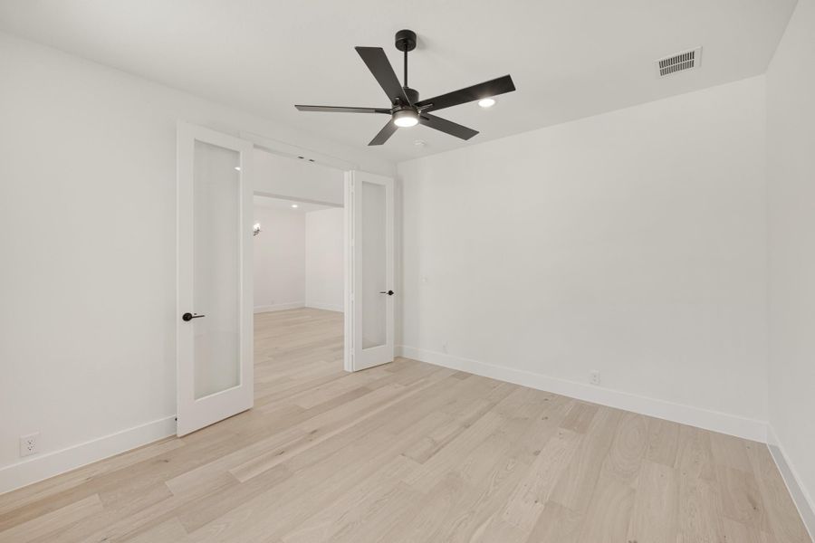 Empty bedroom with light hardwood floors, black ceiling fan, and French doors with glass panels.