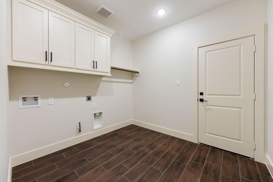 A spacious laundry room with dark wood-like tile flooring. It features upper storage cabinets, a hanging rod, and hookups for a washer and dryer. The room has a clean, modern look with neutral wall colors.