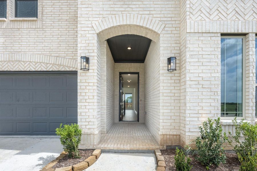 Home entrance with arched brick entryway, black front door, wall sconces, and two-car garage on left Home entrance with arched brick entryway, black front door, wall sconces, and two-car garage on left