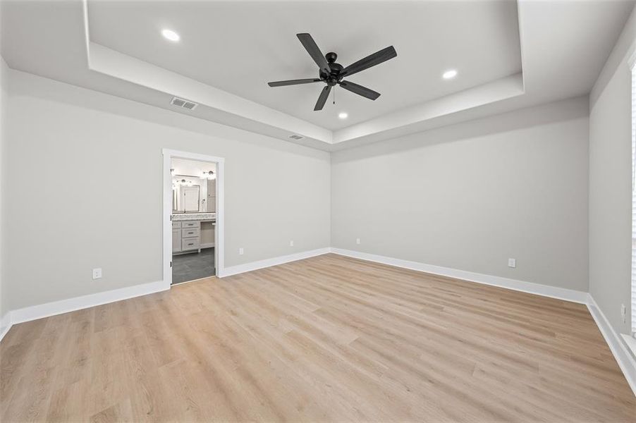 Unfurnished bedroom featuring a raised ceiling, light wood-style floors, a ceiling fan, ensuite bathroom, and recessed lighting Unfurnished bedroom featuring a raised ceiling, light wood-style floors, a ceiling fan, ensuite bathroom, and recessed lighting