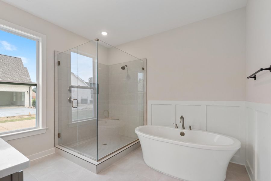 Freestanding soaking tub, glass-enclosed shower with tiled walls, paneled wainscoting, and large window with white trim Freestanding soaking tub, glass-enclosed shower with tiled walls, paneled wainscoting, and large window with white trim