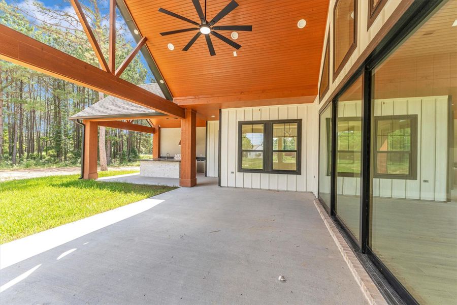 Showcases a spacious covered patio with a high wooden ceiling and ceiling fan, offering a seamless indoor-outdoor connection. Large glass doors and windows provide ample natural light, and the area overlooks a lush, green lawn surrounded by trees, ideal for relaxation or entertaining.