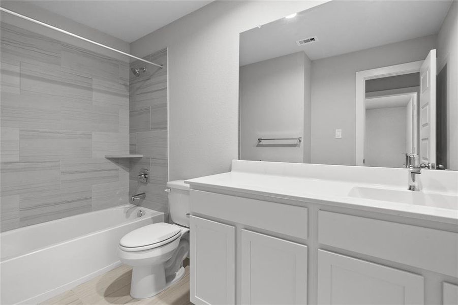 Secondary bath features tile flooring, bath/shower combo with tile surround, stained wood cabinets, beautiful light countertops, mirror, dark, sleek fixtures and modern finishes.