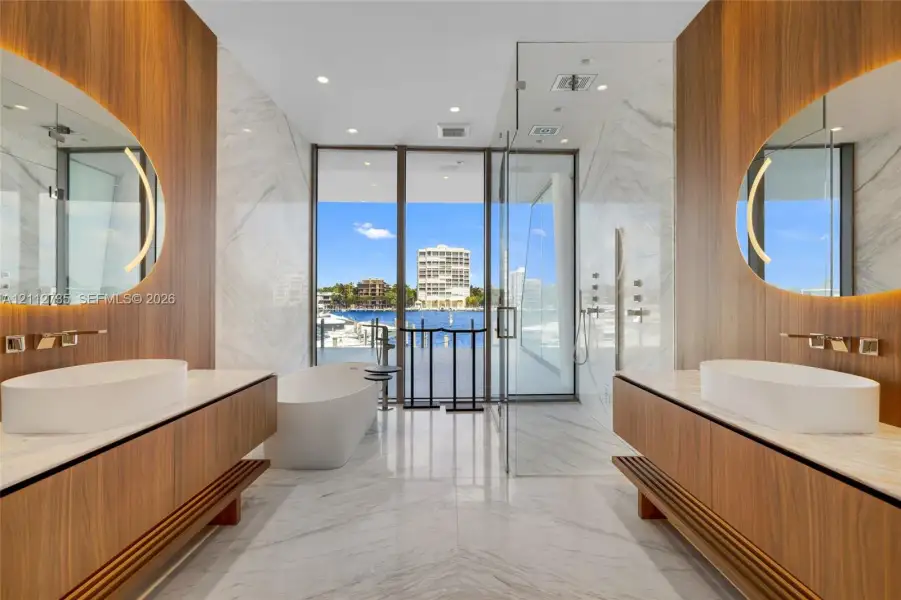 Dual floating wood-finish vanities with vessel sinks and integrated backlit mirrors, freestanding soaking tub, expansive stone-tiled floor, and a glass-enclosed walk-in shower